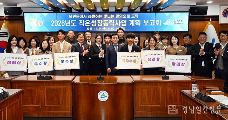 밀양시청 소회의실에서 열린 2026년도 작은성장동력사업 계획 보고회에서, 평가 결과 선정된 읍·면·동 직원들이 사업 선정 표찰을 들고 단체 사진을 촬영하고 있다.